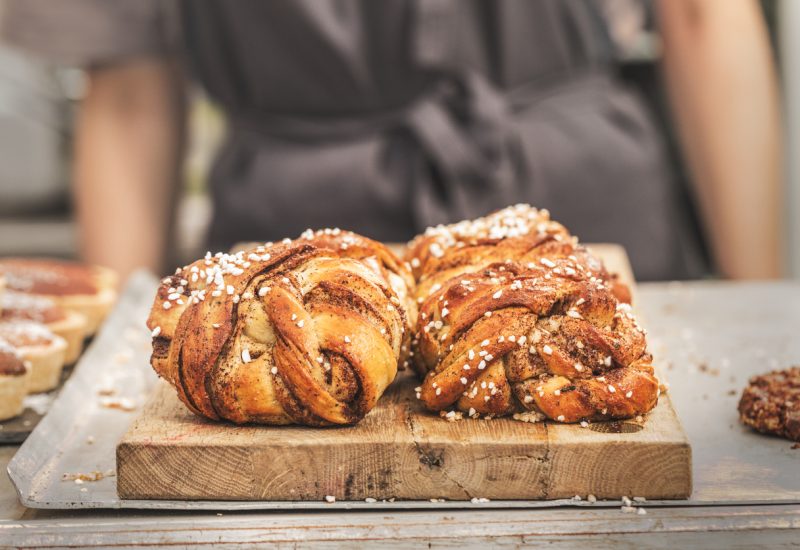 Twisted traditional Swedish cinnamon buns at a café. The sweet buns are on a wooden chopping board, and there is an  unrecognizable person with an apron in the background.